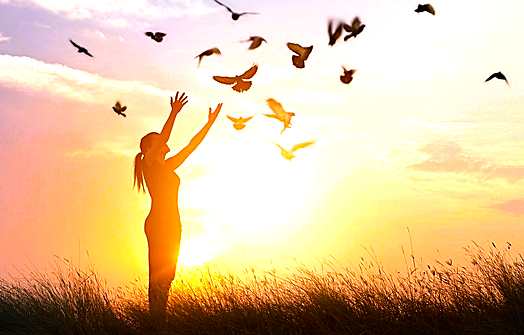 Femme heureuse libérée d'un envoutement qui relâche des oiseaux devant un couché de soleil.